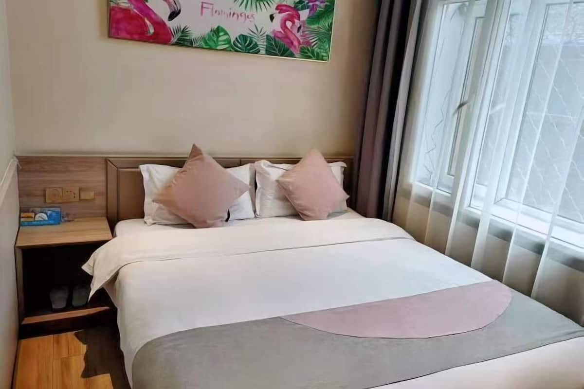 Uma cama bem arrumada com esquema de cores branco e rosa, um criado-mudo com abajur e uma janela com cortinas.