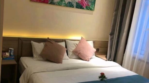 A hotel room with a bed, pillows, a nightstand, a painting on the wall, and a window with curtains.