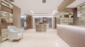 A modern hotel lobby with a reception desk, seating area, and shelving with items.