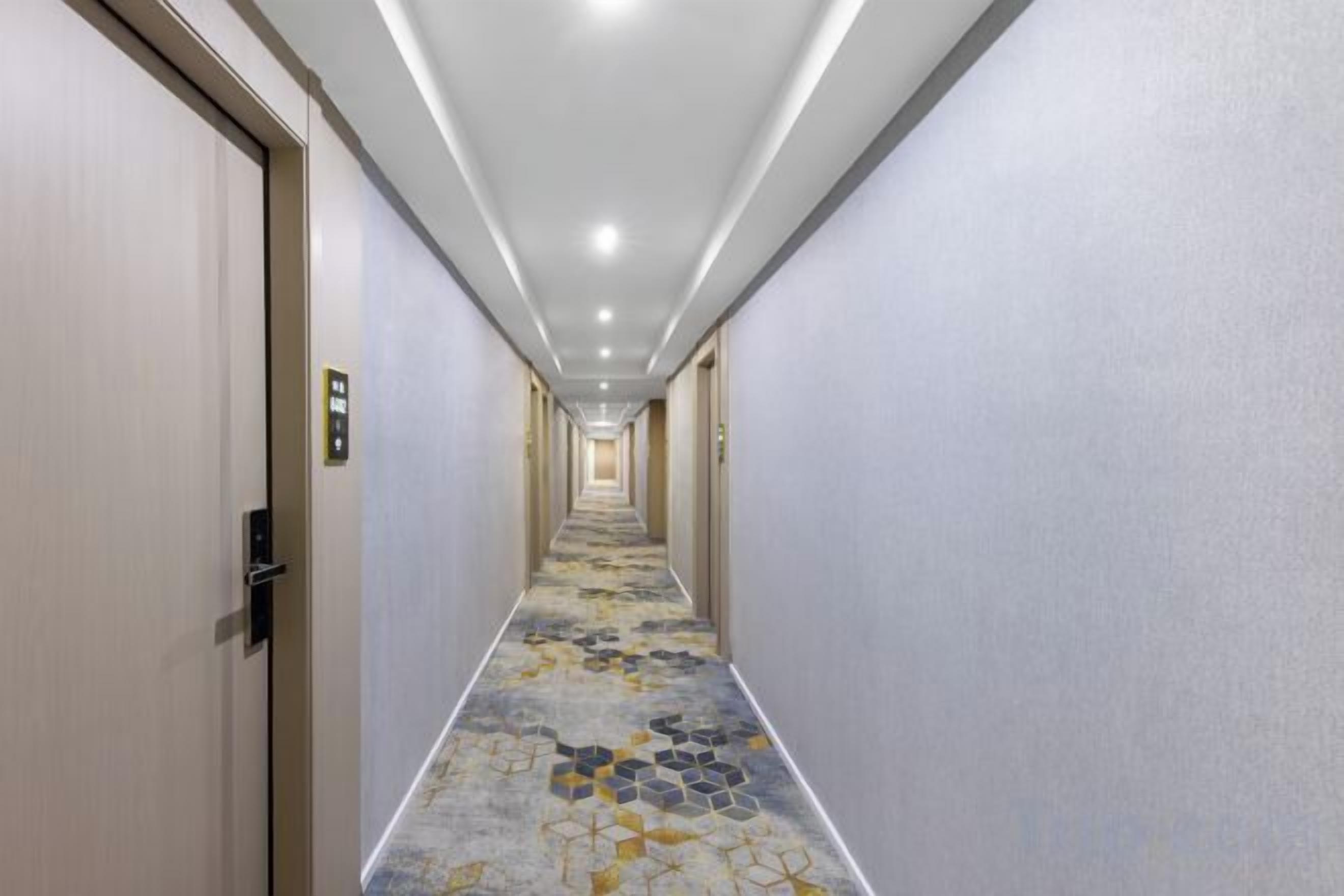 A hotel corridor with doors on both sides, a patterned carpet, and a ceiling with recessed lighting.