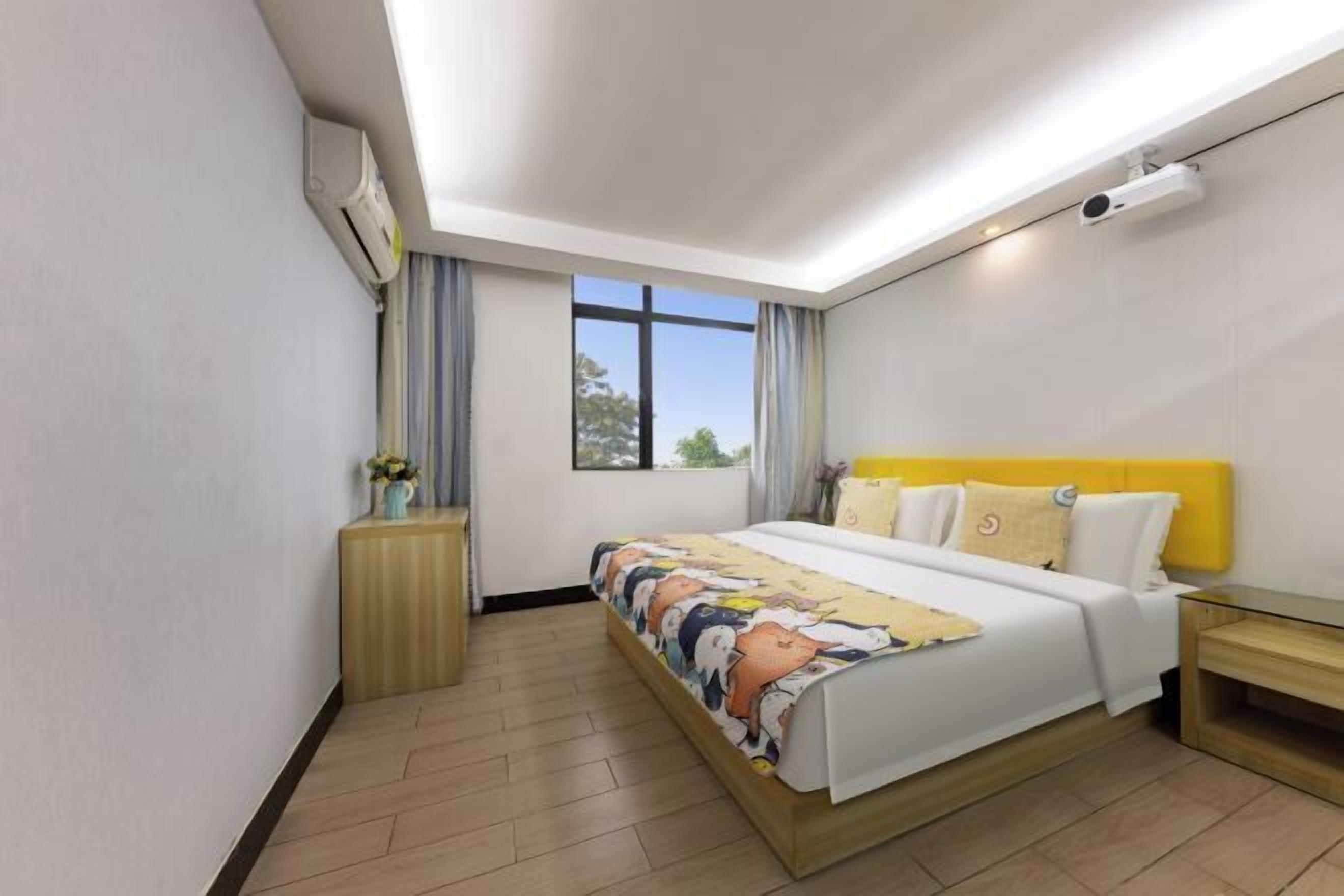 A modern hotel room with a large bed, a bedside table, a nightstand, a window with curtains, and a ceiling-mounted projector.