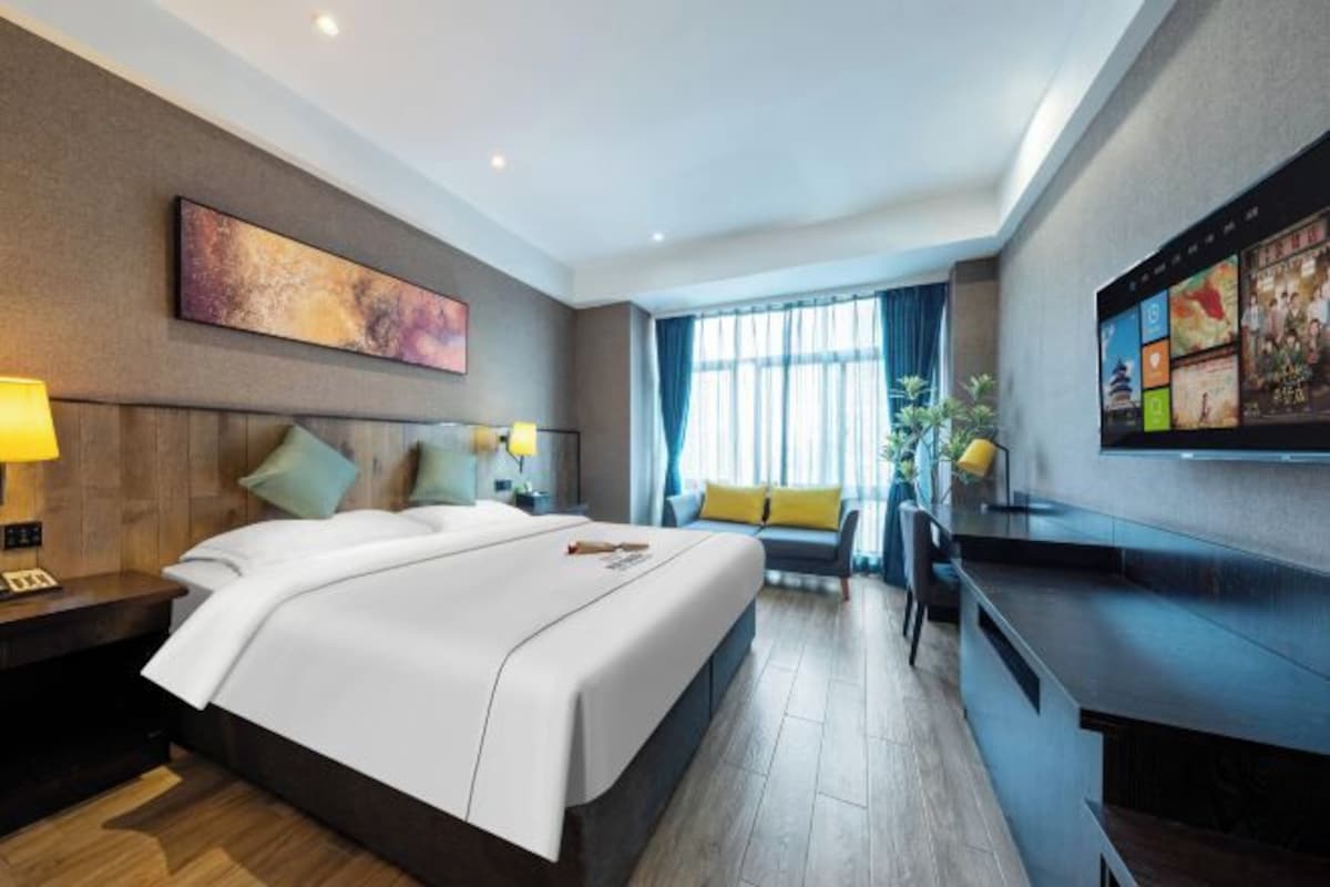 A hotel room with a bed, bedside table, lamp, artwork, window, and a TV.