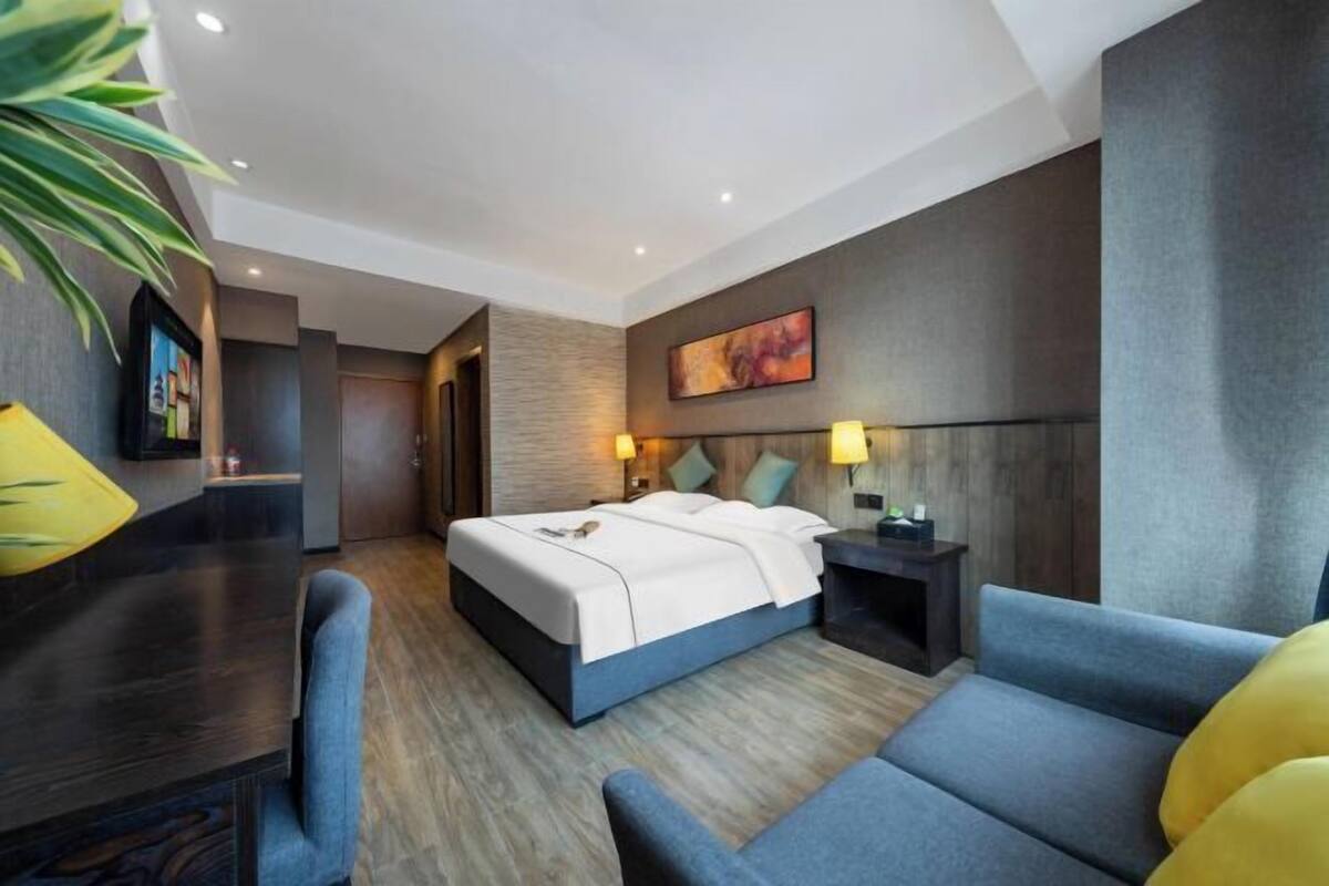 A modern hotel room with a bed, a desk, a chair, a sofa, and a painting on the wall.