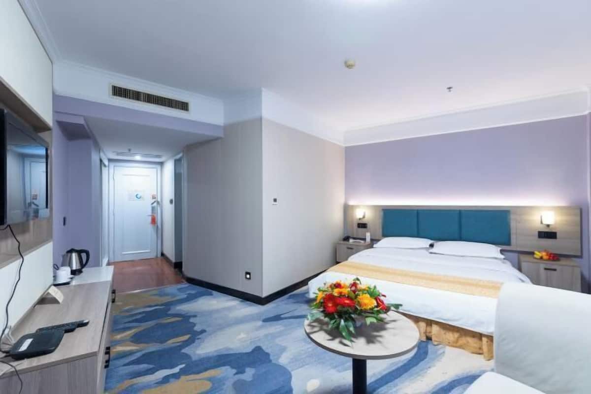A hotel room with a bed, bedside tables, a desk with a chair, a small round table with a flower arrangement, and a view of a corridor.