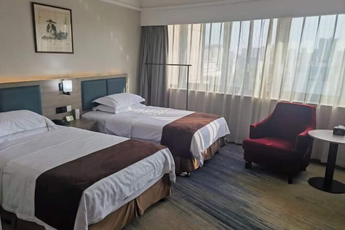 A hotel room with two beds, a red armchair, a small round table, and a large window with curtains.
