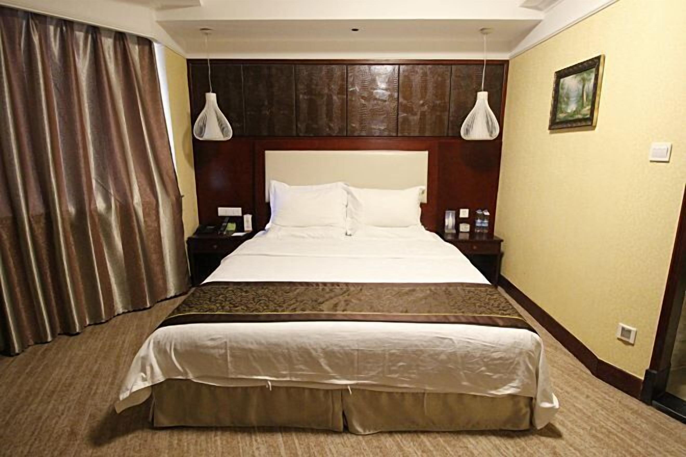 Habitación de hotel con una cama grande, dos mesitas de noche, una mesita auxiliar, cabecero y un cuadro en la pared, además de una cortina.