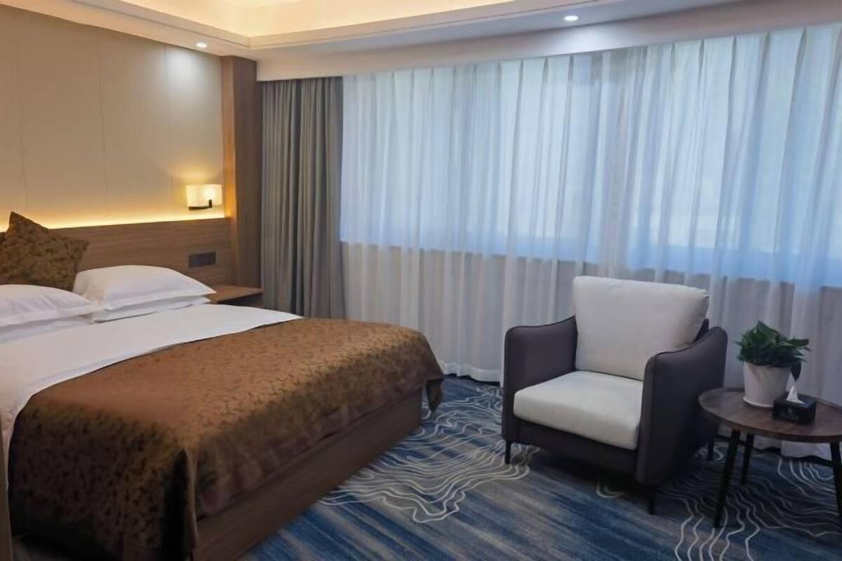 A hotel room with a bed, a chair, a small table with a plant, and a window with curtains.