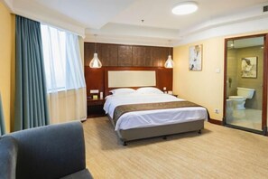Habitación de hotel con cama, una silla, una mesita de noche, una lámpara y un baño visible a través de una puerta de cristal.