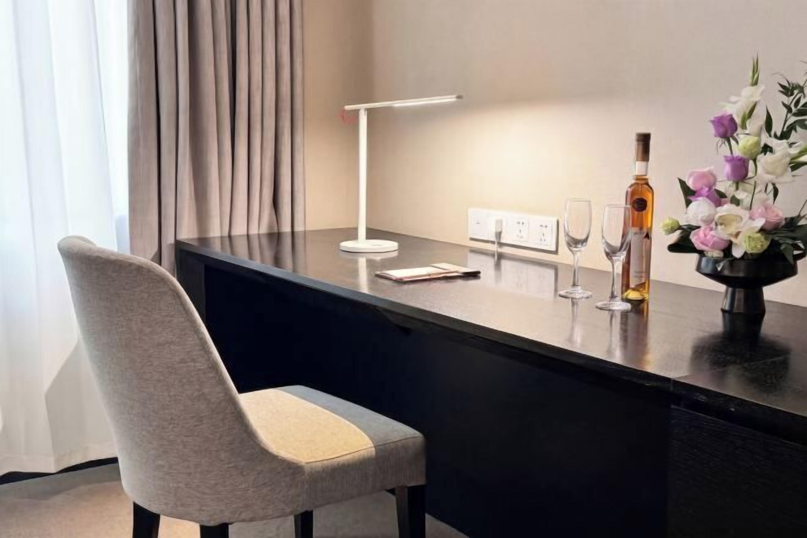Ein modernes Hotelzimmer mit Schreibtisch, Lampe, Blumen in einer Vase und einer Weinflasche.
