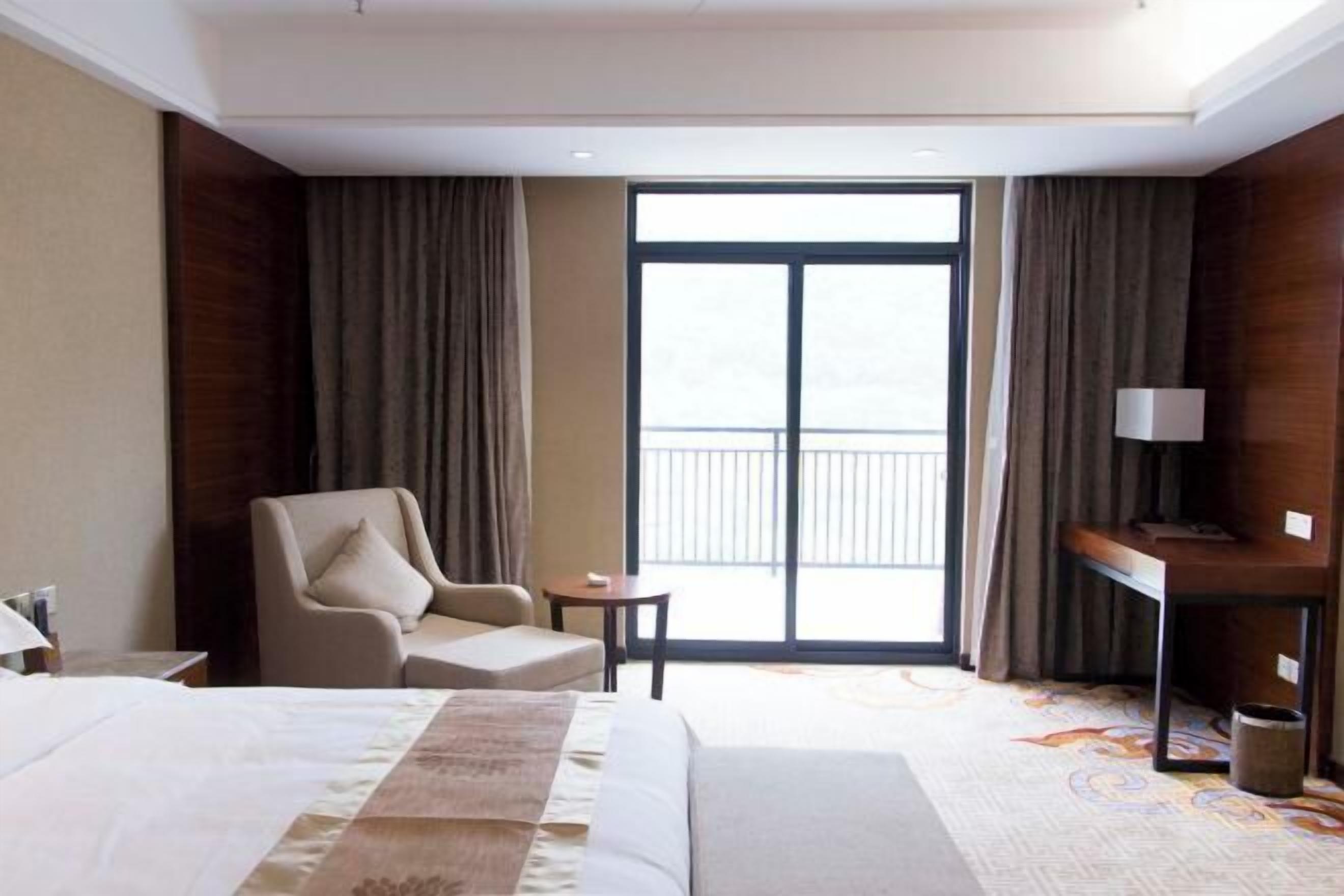 A hotel room with a bed, a chair, a desk, a lamp, and a large window with a view.