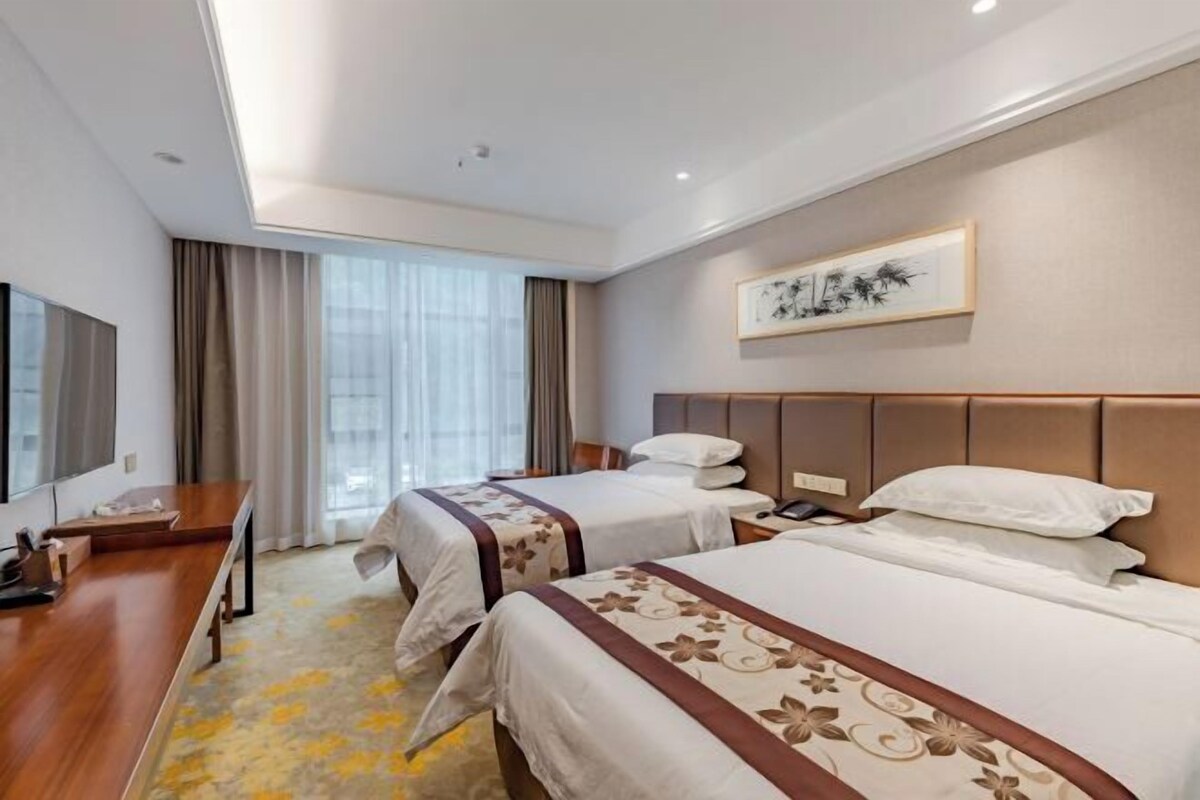 A hotel room with two beds, a desk, a television, and a large window with curtains.