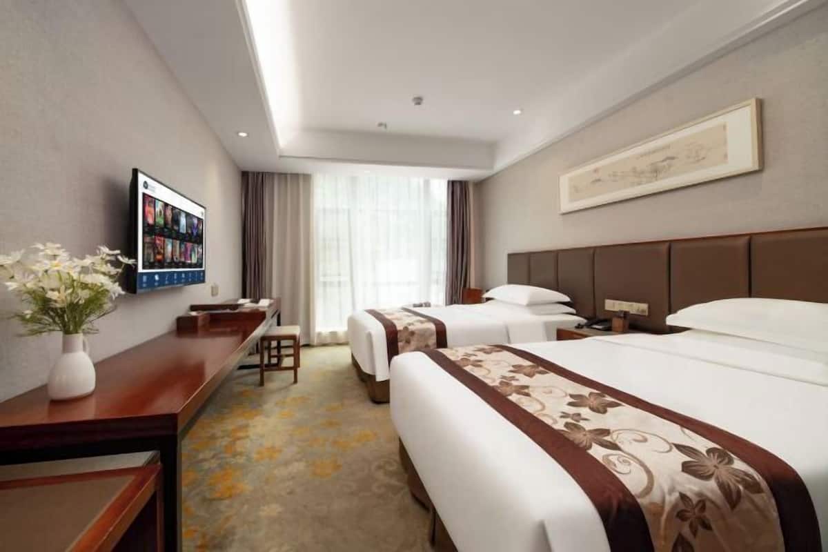 A hotel room with two beds, a desk, a chair, a TV, and a large window with curtains.