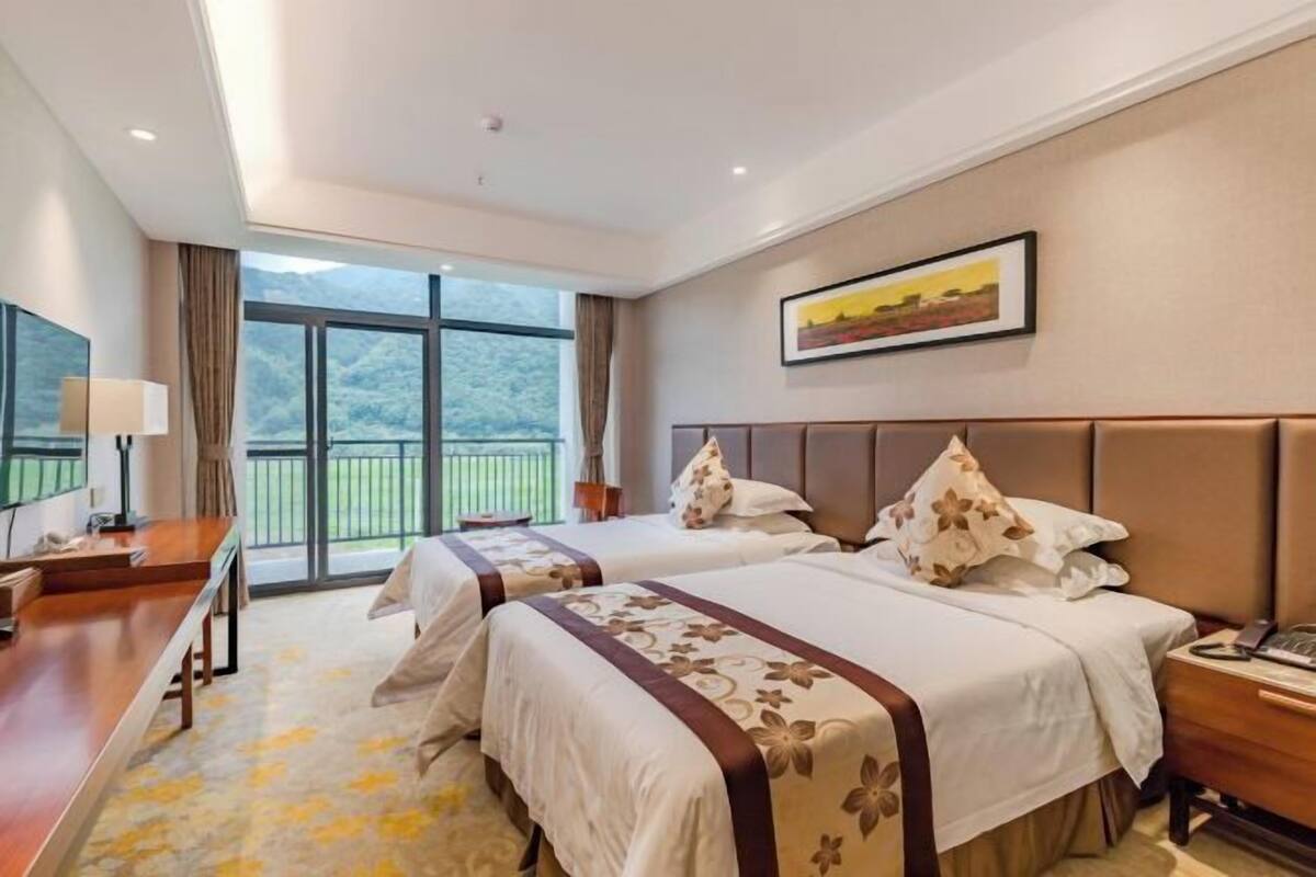 Ein Hotelzimmer mit zwei Betten, einem Schreibtisch, einem Stuhl, einem großen Fenster mit Blick und einem Bild an der Wand.