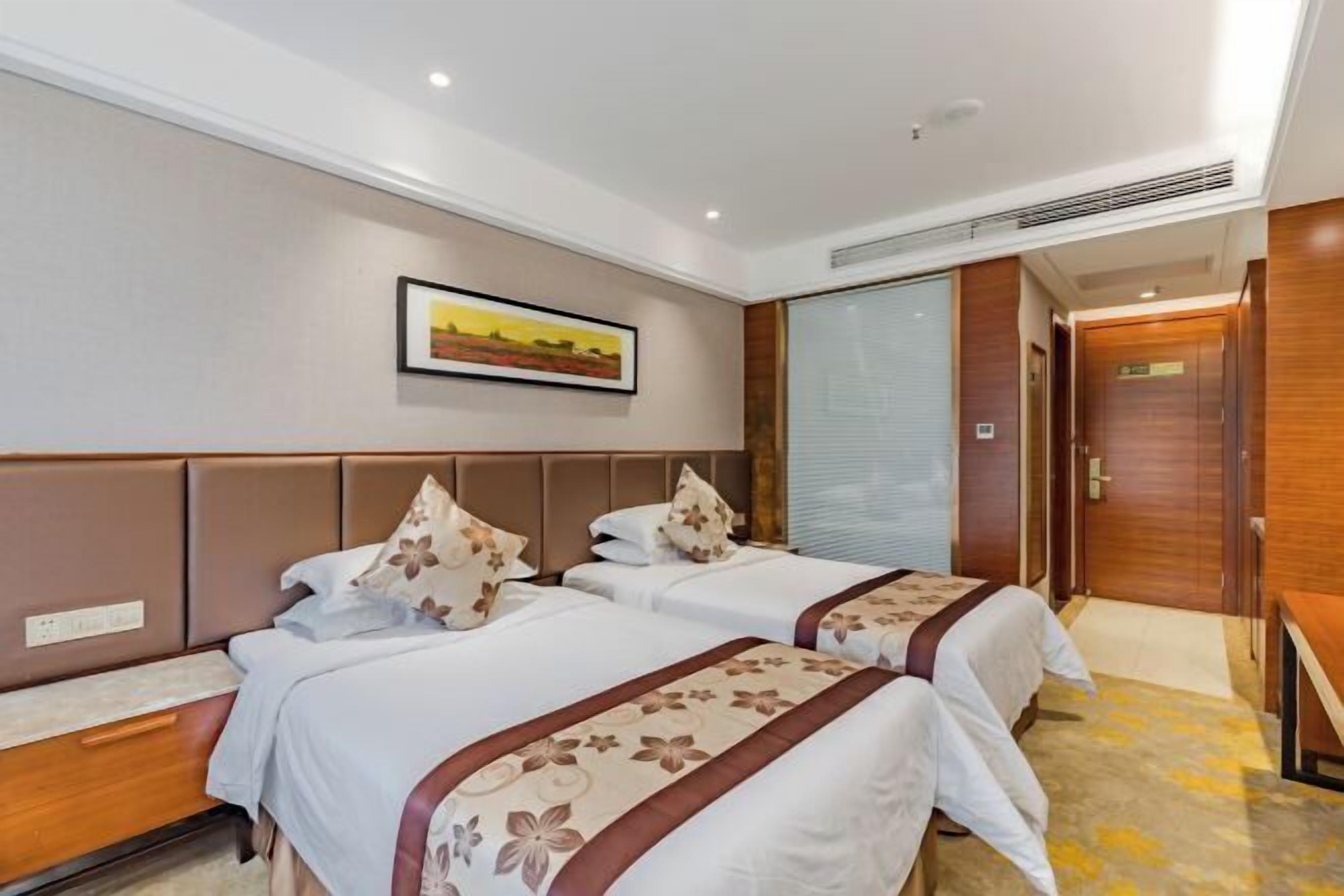 A hotel room with two beds, a wooden headboard, a painting on the wall, and a door with a wooden panel.