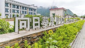 Un edificio con un cartel en primer plano, rodeado de vegetación y montañas al fondo.