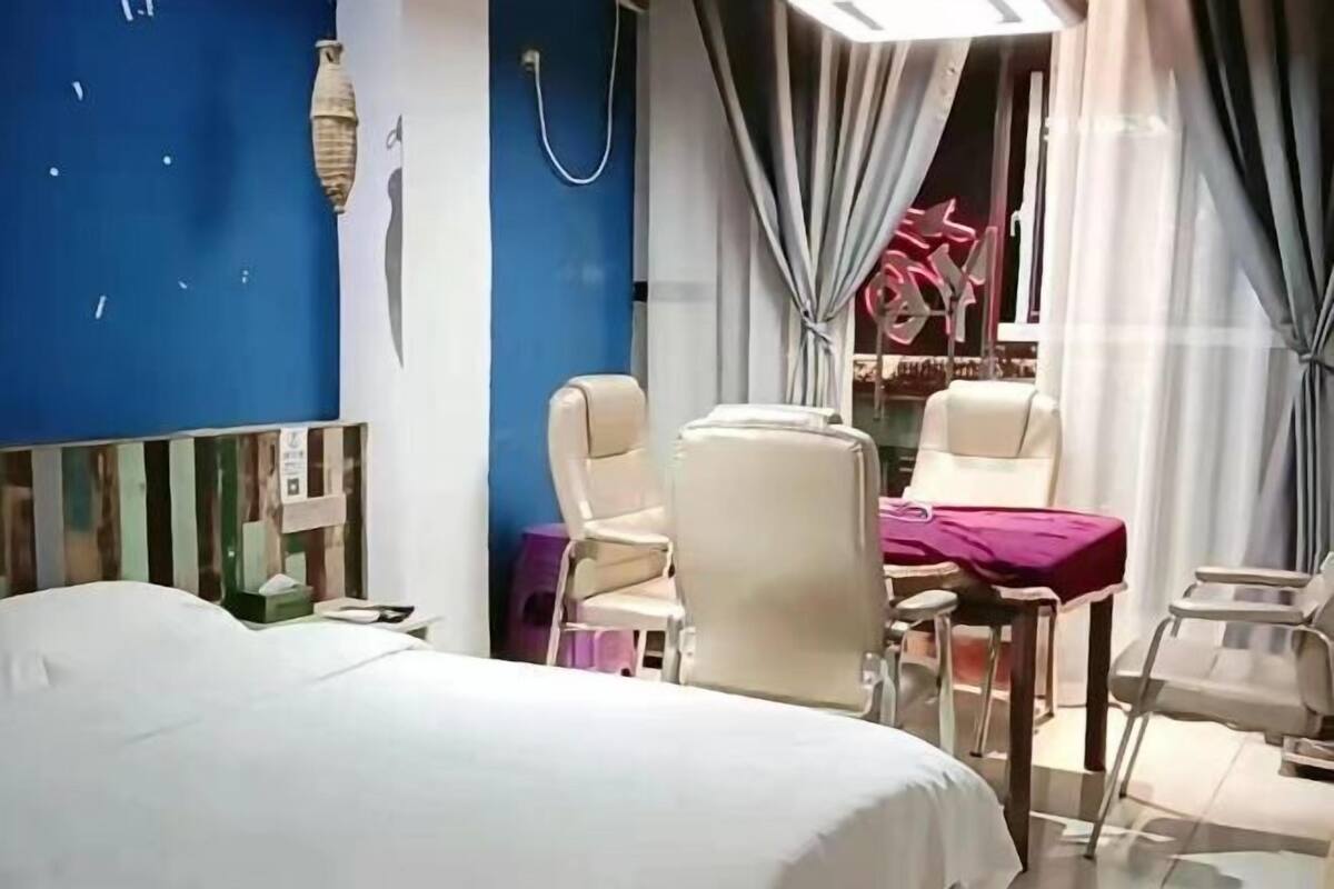 A hotel room with a bed, two chairs, a small table with a purple tablecloth, a vanity with a mirror, and a blue wall with a decorative item.