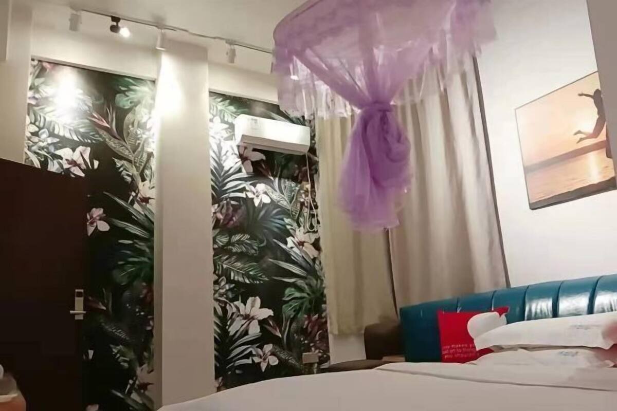 A hotel room with a bed, a window with curtains, a wall with a tropical wallpaper, and a ceiling light.