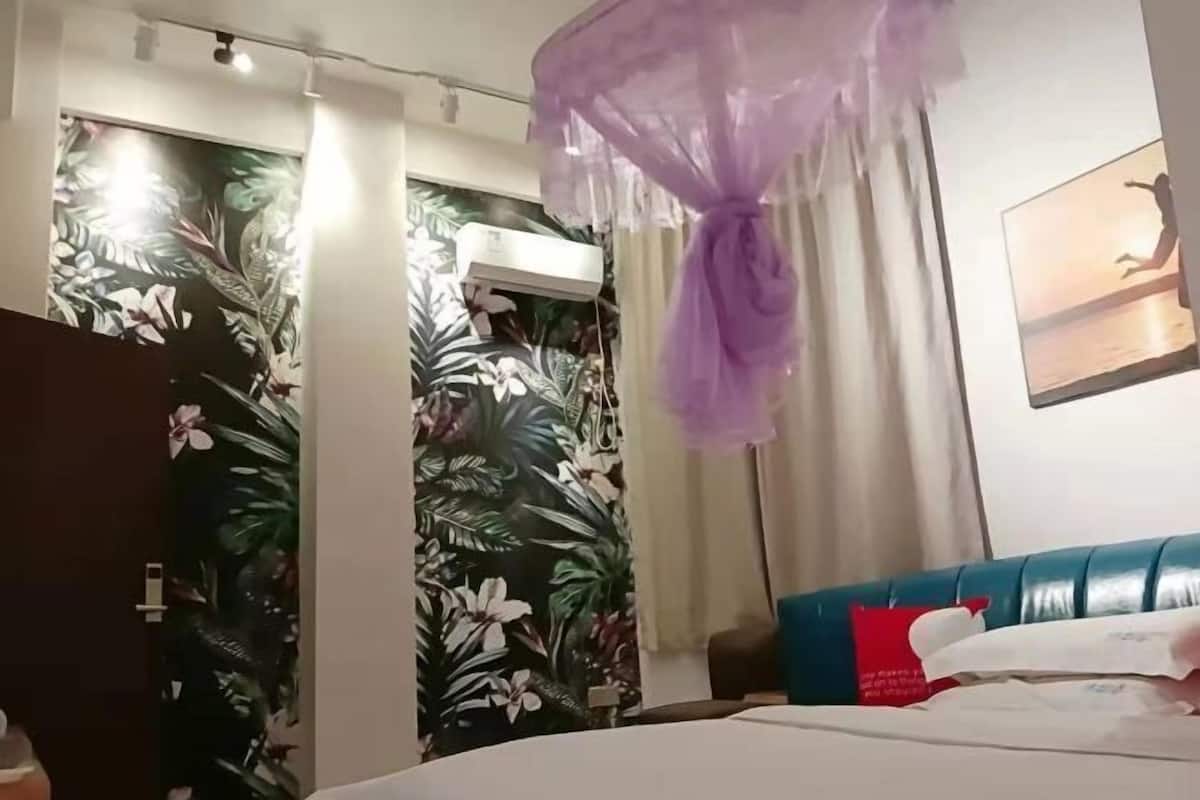 A hotel room with a bed, a window with curtains, a wall with a tropical wallpaper, and a ceiling light.