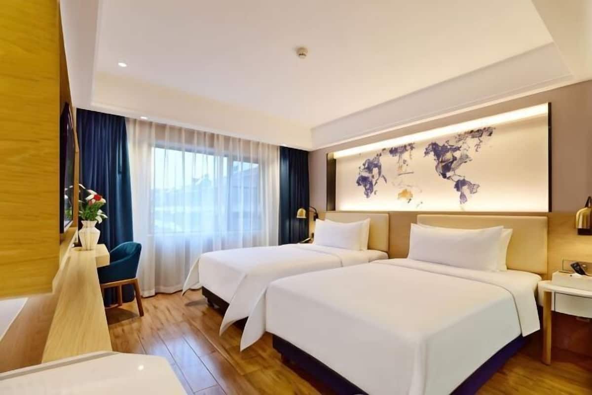 A hotel room with two beds, a desk, a chair, a world map on the wall, and a large window with curtains.