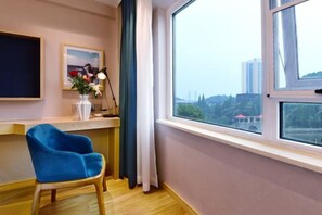 Een kamer met een groot raam, een blauwe stoel, een witte vaas met bloemen en een lijst met een afbeelding aan de muur.