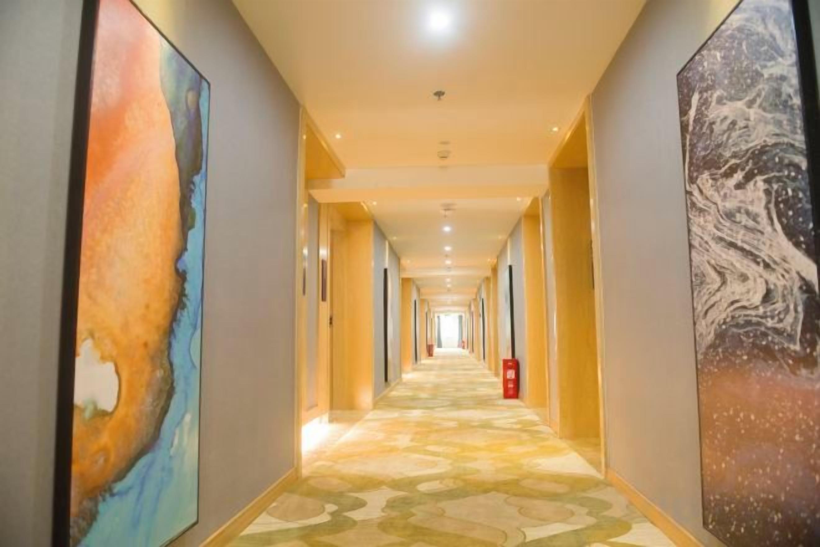 A hotel hallway with a patterned carpet, framed artwork on the walls, and a fire extinguisher.