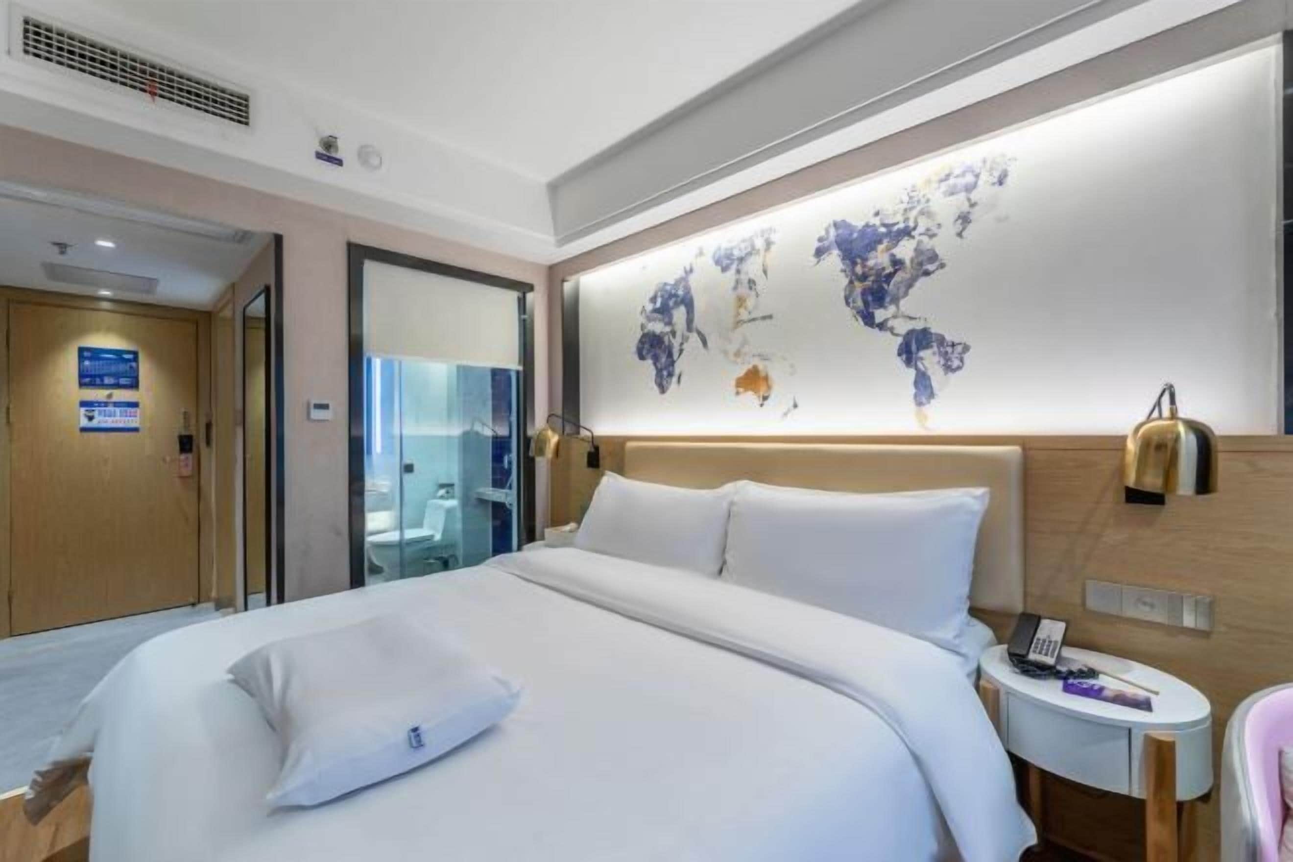 A modern hotel room with a large bed, a bedside table with a lamp, a world map on the wall, and a bathroom visible through a glass door.