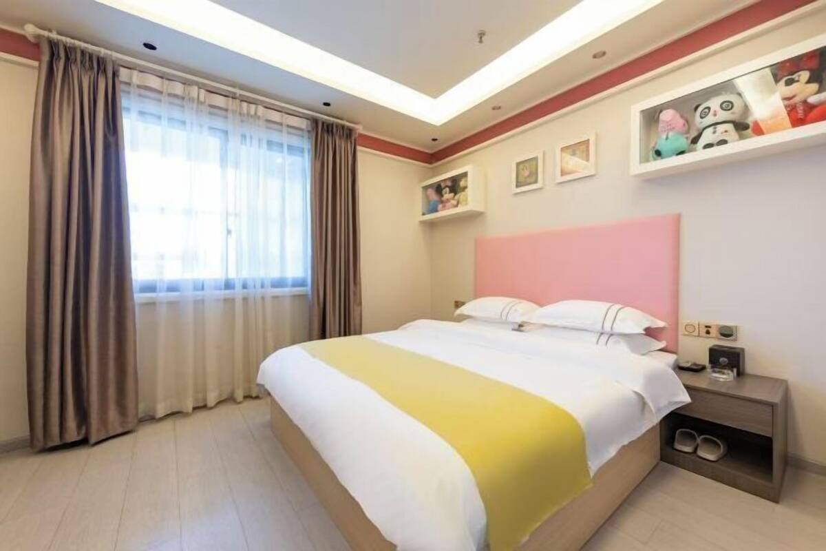 Habitación de hotel con cama, cortinas, mesita de noche con pantuflas, estante con peluches y estante de pared con fotos enmarcadas.