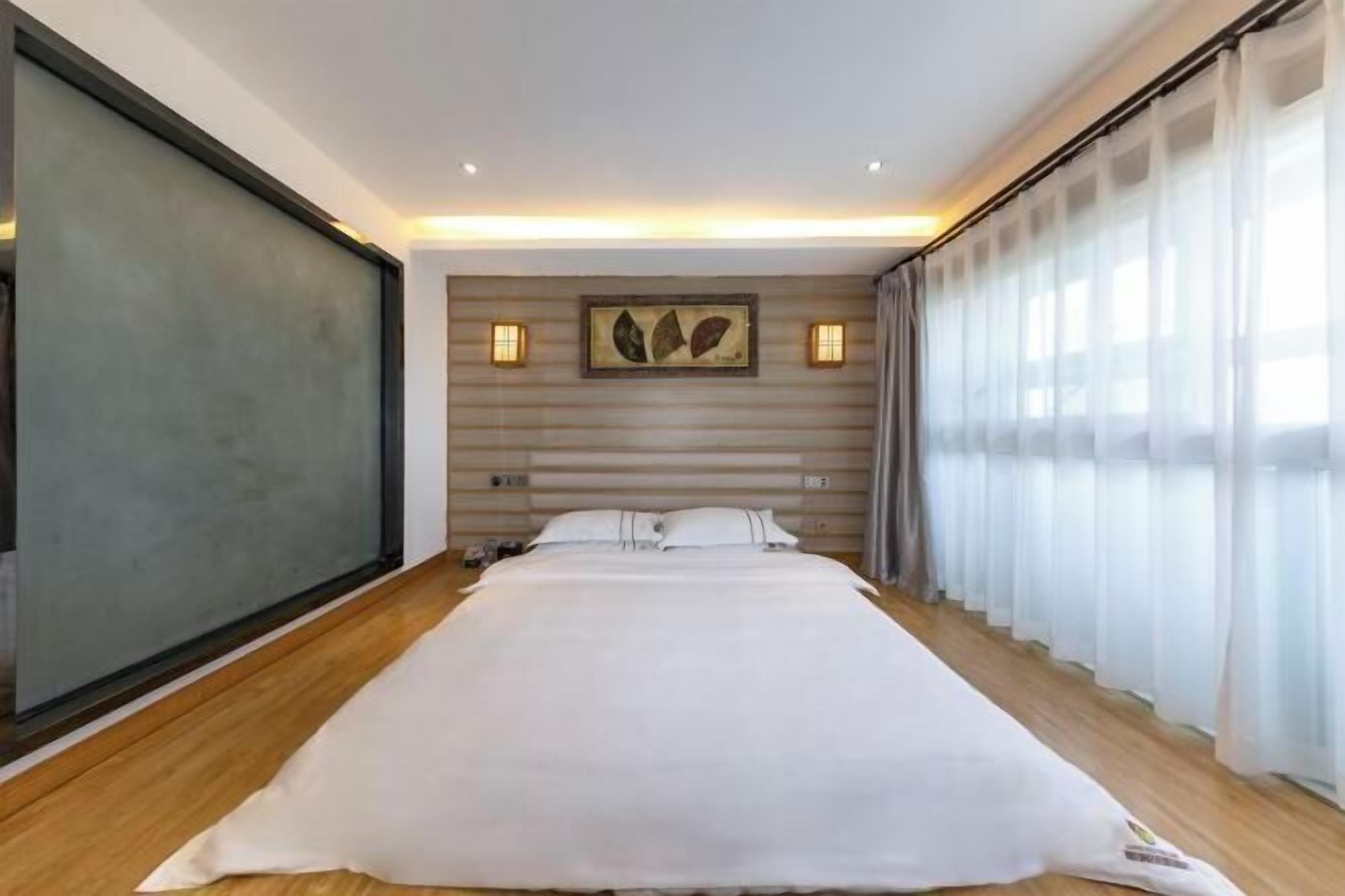 A modern hotel room with a large bed, a headboard with artwork, two bedside lamps, and a large window with sheer curtains.