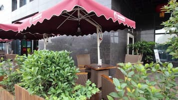 Un área de estar exterior con un paraguas rojo, mobiliario de mimbre y plantas en macetas.