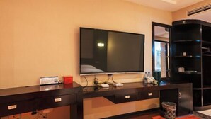 Ein Hotelzimmer mit einem großen Flachbildfernseher an der Wand, einem dunklen Holztisch mit Schubladen, einer kleinen Mülleimer und einem schwarzen Regal.