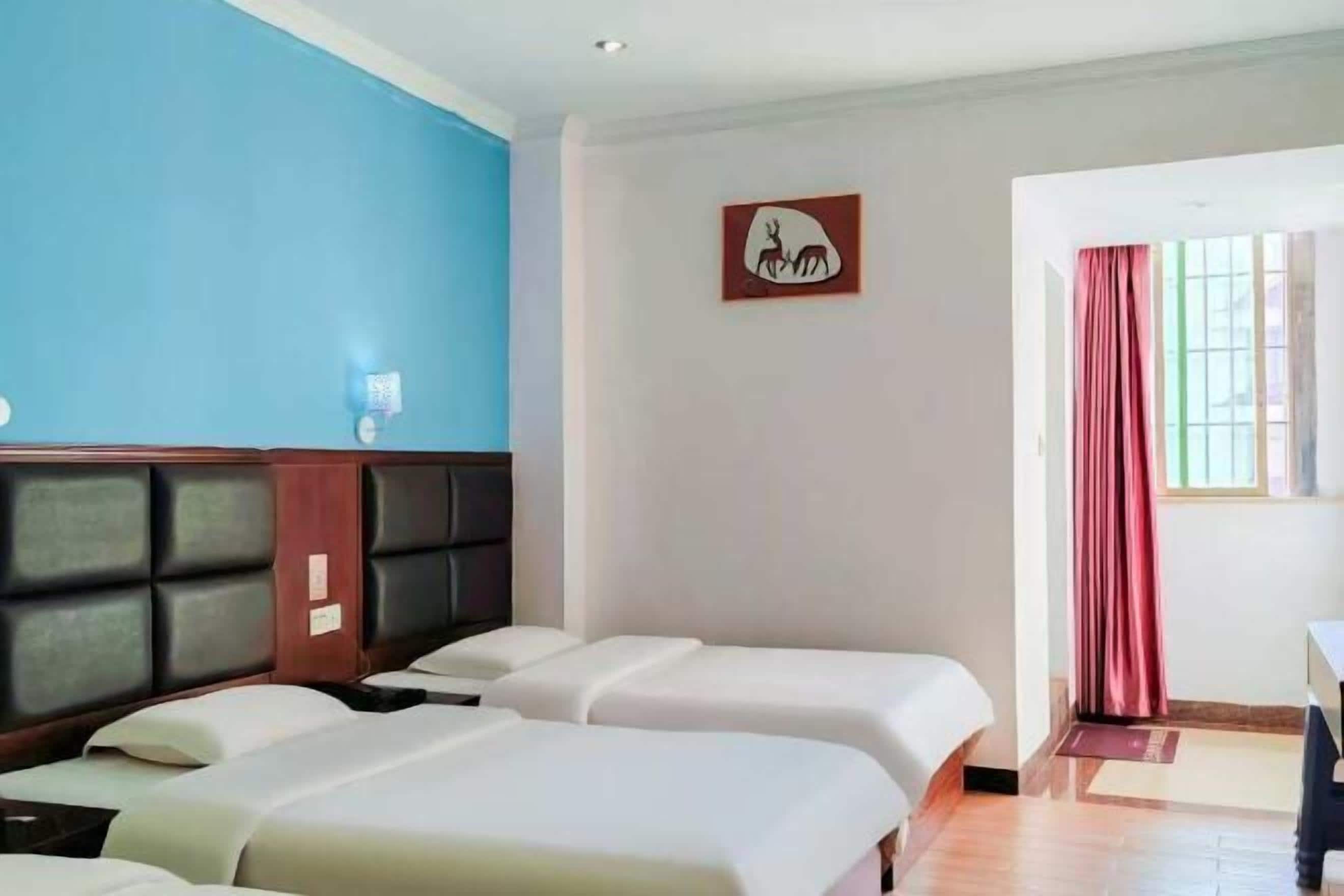 A hotel room with two beds, a blue wall, a wooden headboard, a framed picture, a window with curtains, and a small table.