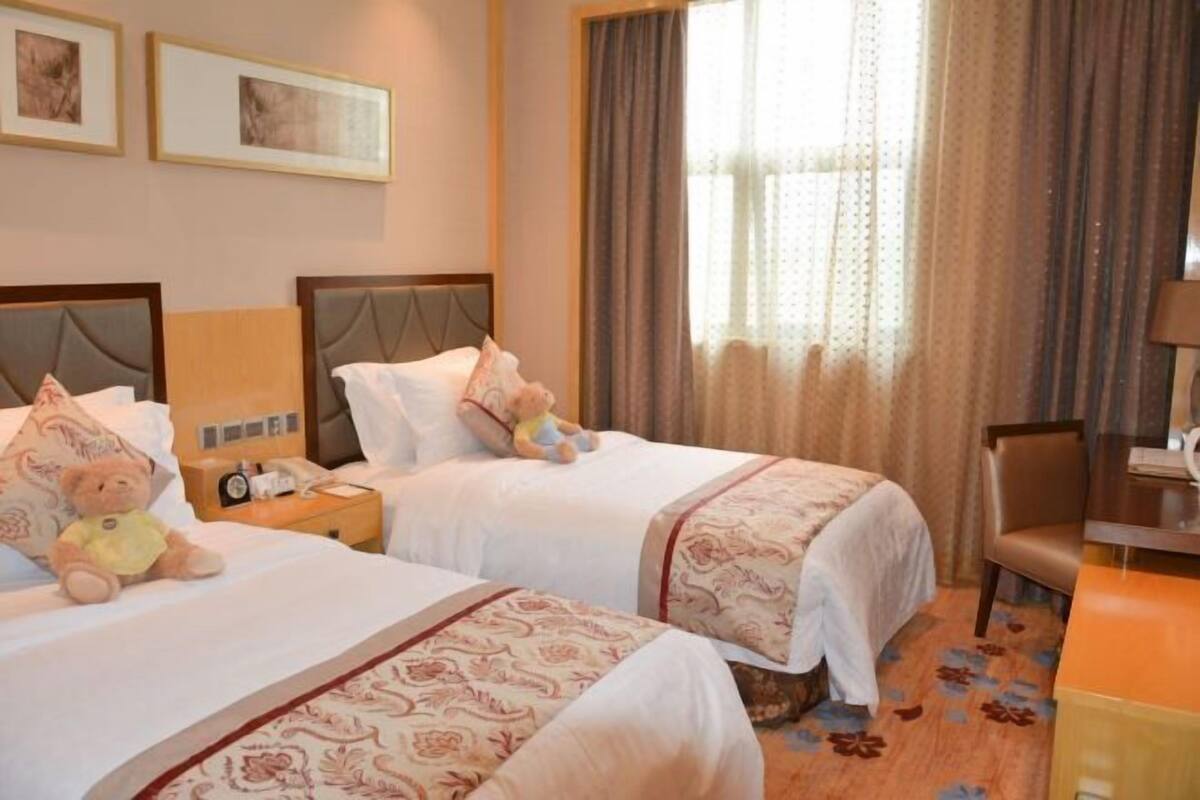Une chambre d’hôtel avec deux lits, un bureau et une chaise. Les murs sont ornés de tableaux encadrés et il y a une fenêtre avec des rideaux.