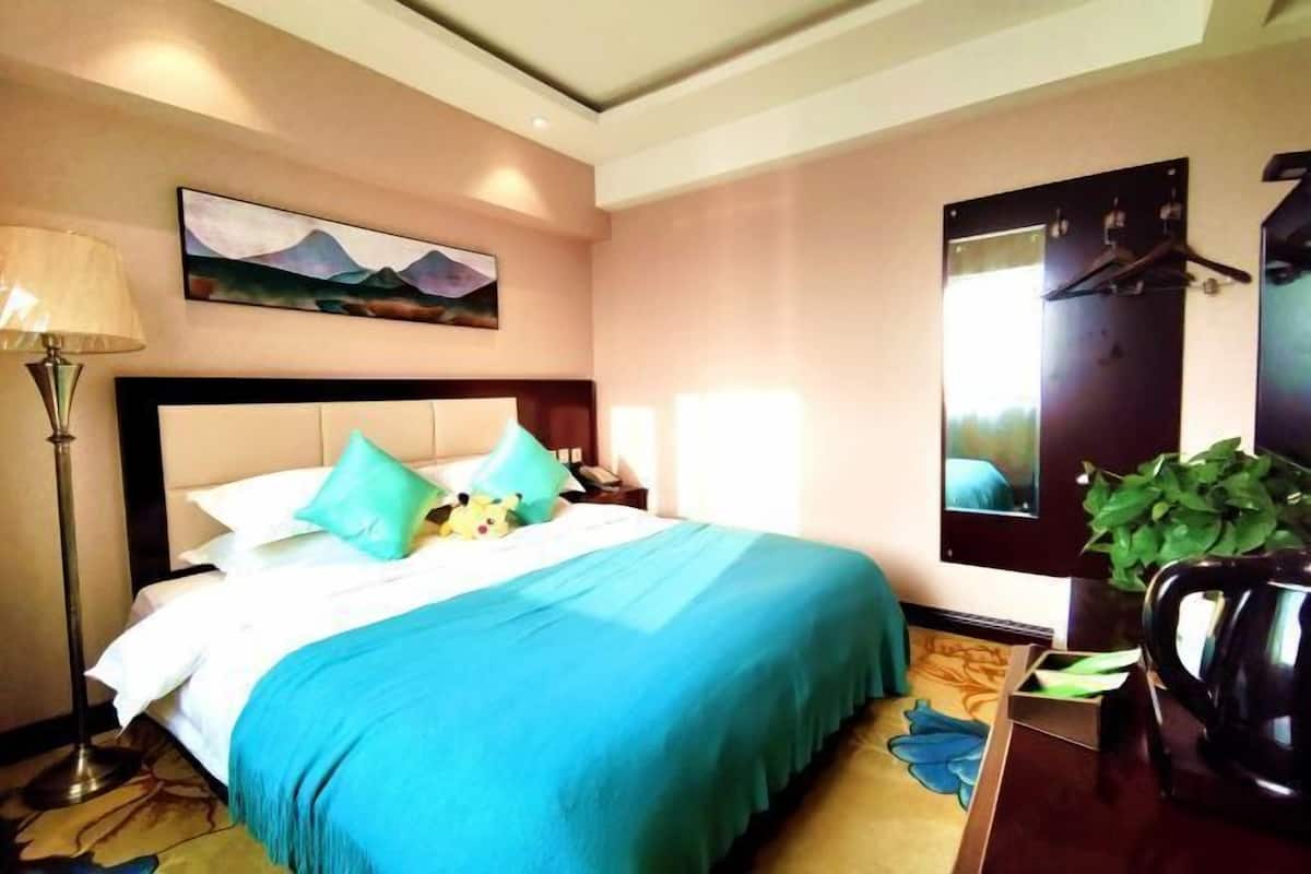 A hotel room with a bed, a nightstand, a lamp, a painting, a mirror, and a plant.
