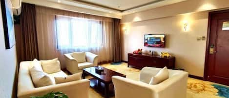Habitación de hotel con un sofá, televisor, una mesita de noche de madera oscura y un ventanal con cortinas.