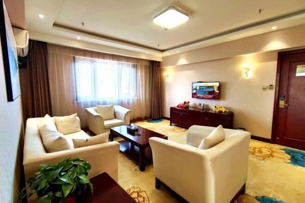 A hotel room with a sofa set, a television, a dark wooden side table, and a large window with curtains.