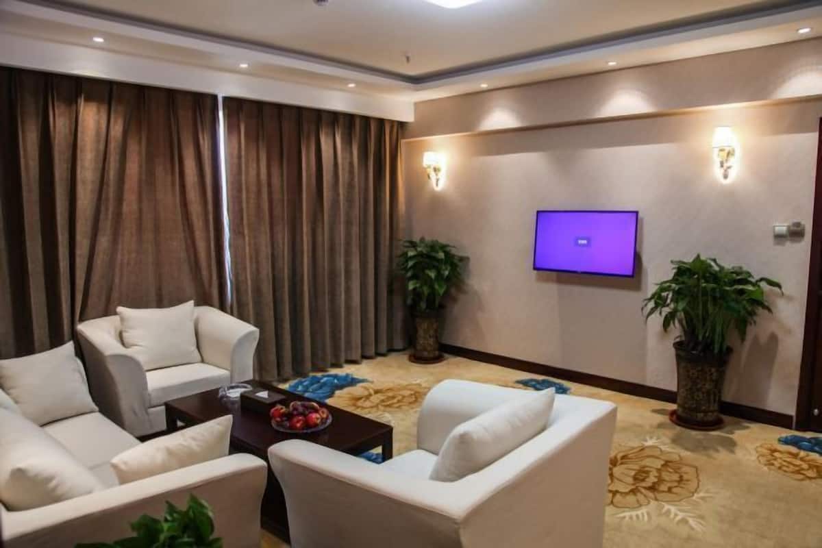 A hotel room with a flat-screen TV mounted on the wall, a sofa, two armchairs, a coffee table, and two potted plants.