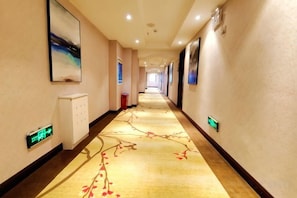 Corredor de hotel com piso coberto por carpete, quadros nas paredes e um relógio digital.