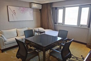 A hotel room with a bed, a white sofa, a dining table with chairs, and a large window with curtains.