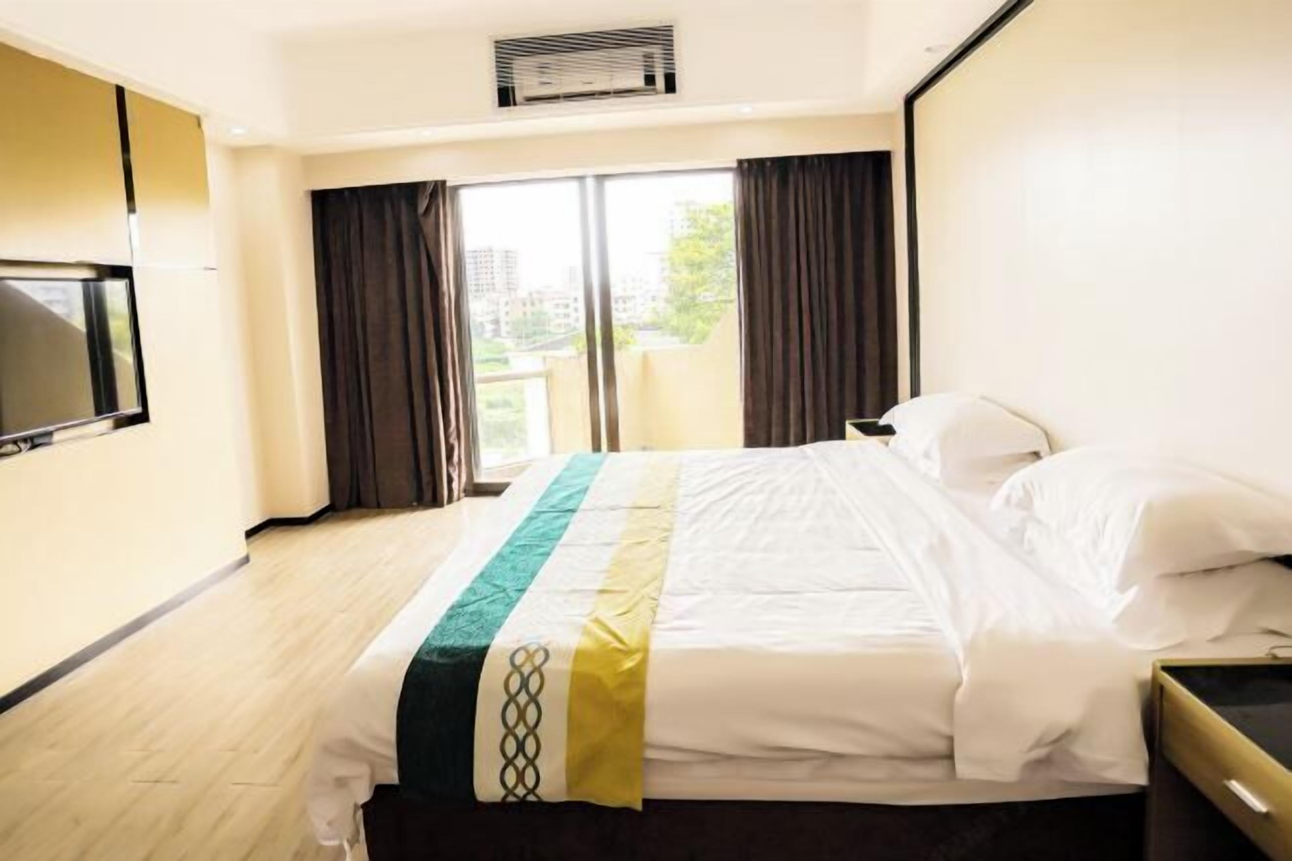 A hotel room with a bed, a TV, a window with curtains, and an air conditioning unit.