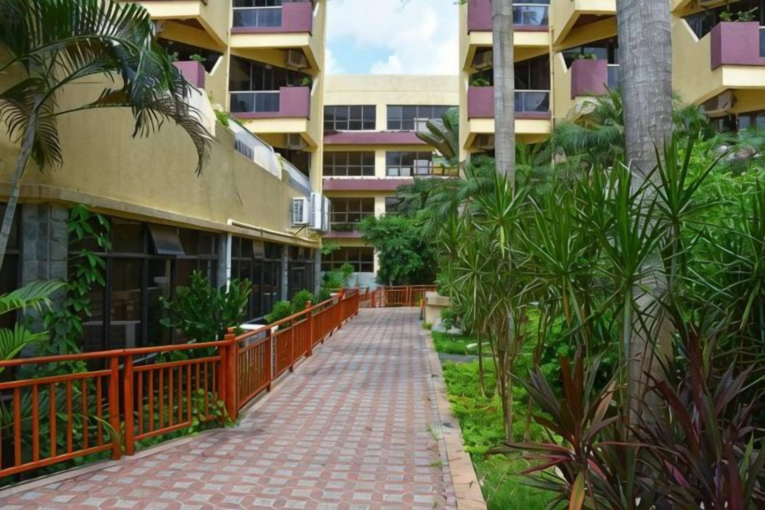 Un sendero conduce a un edificio de varios pisos con balcones, rodeado de vegetación y una cerca roja.