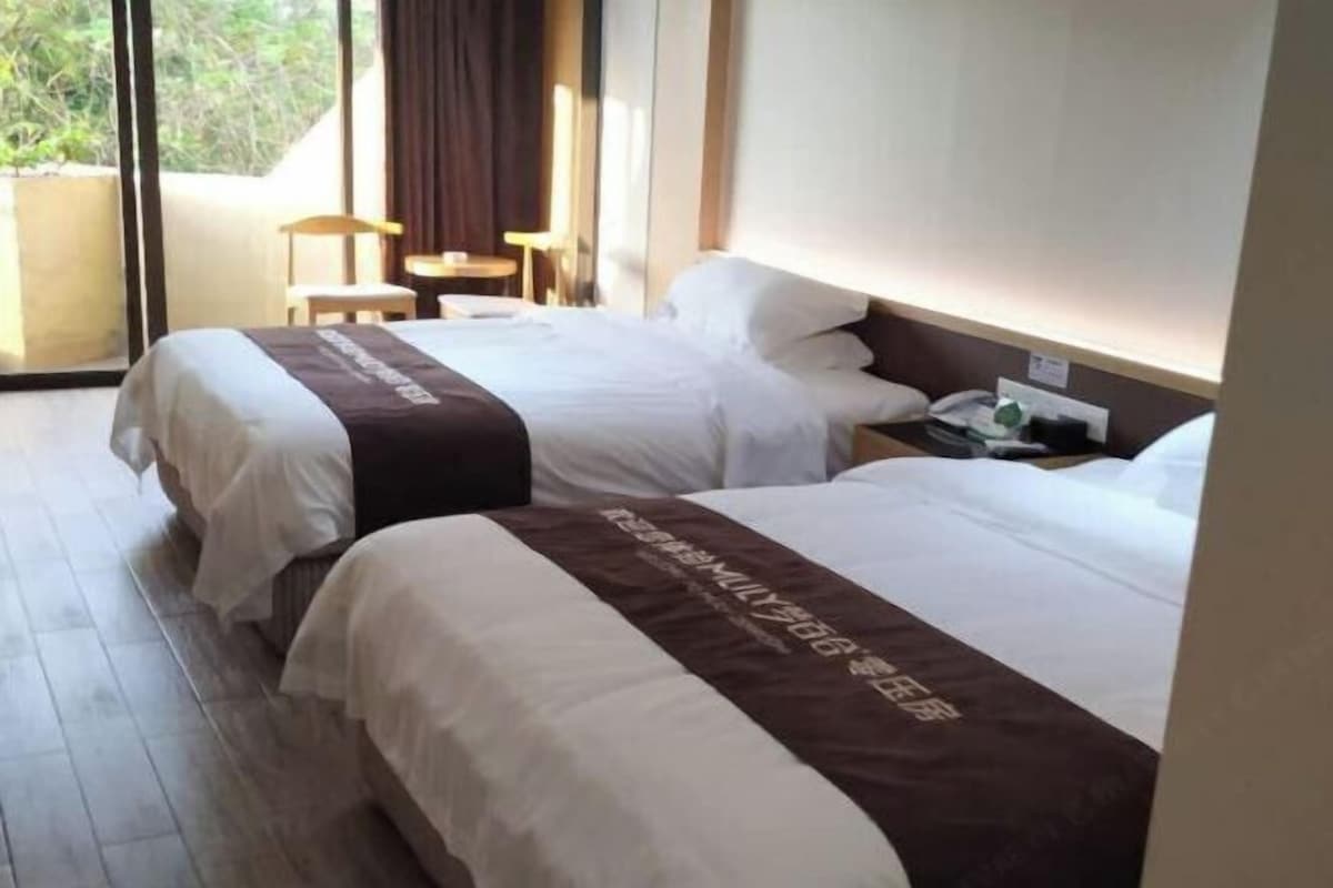 A hotel room with two beds, a small table, a chair, and a window with a view of greenery.