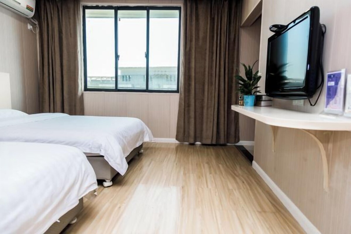 Une chambre d’hôtel avec deux lits, une grande fenêtre avec des rideaux, un téléviseur fixé au mur et une petite étagère avec une plante.