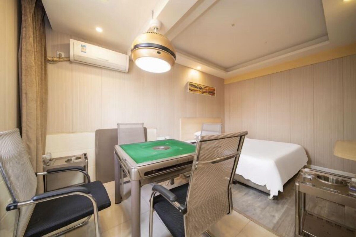 A hotel room with a bed, a desk with a green table, two chairs, a lamp, and an air conditioning unit.