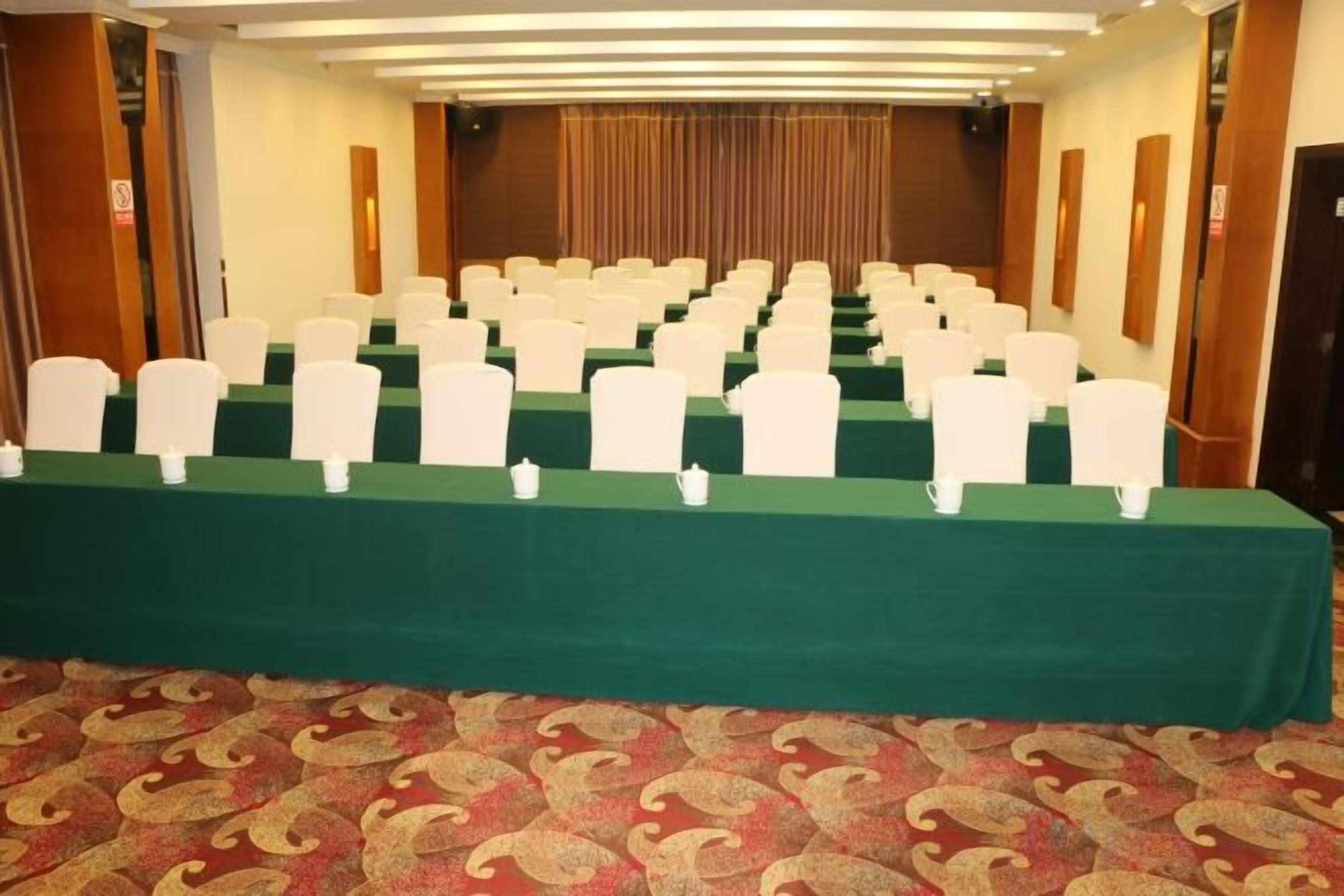 Una sala conferenze con file di sedie bianche e un tavolo verde, pavimento rivestito di moquette e pannelli di legno alle pareti.