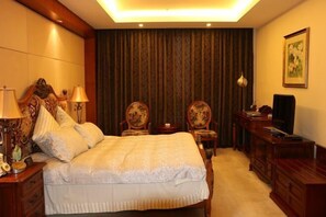 A hotel room with a large bed, two chairs, a desk with a television, and a painting on the wall.
