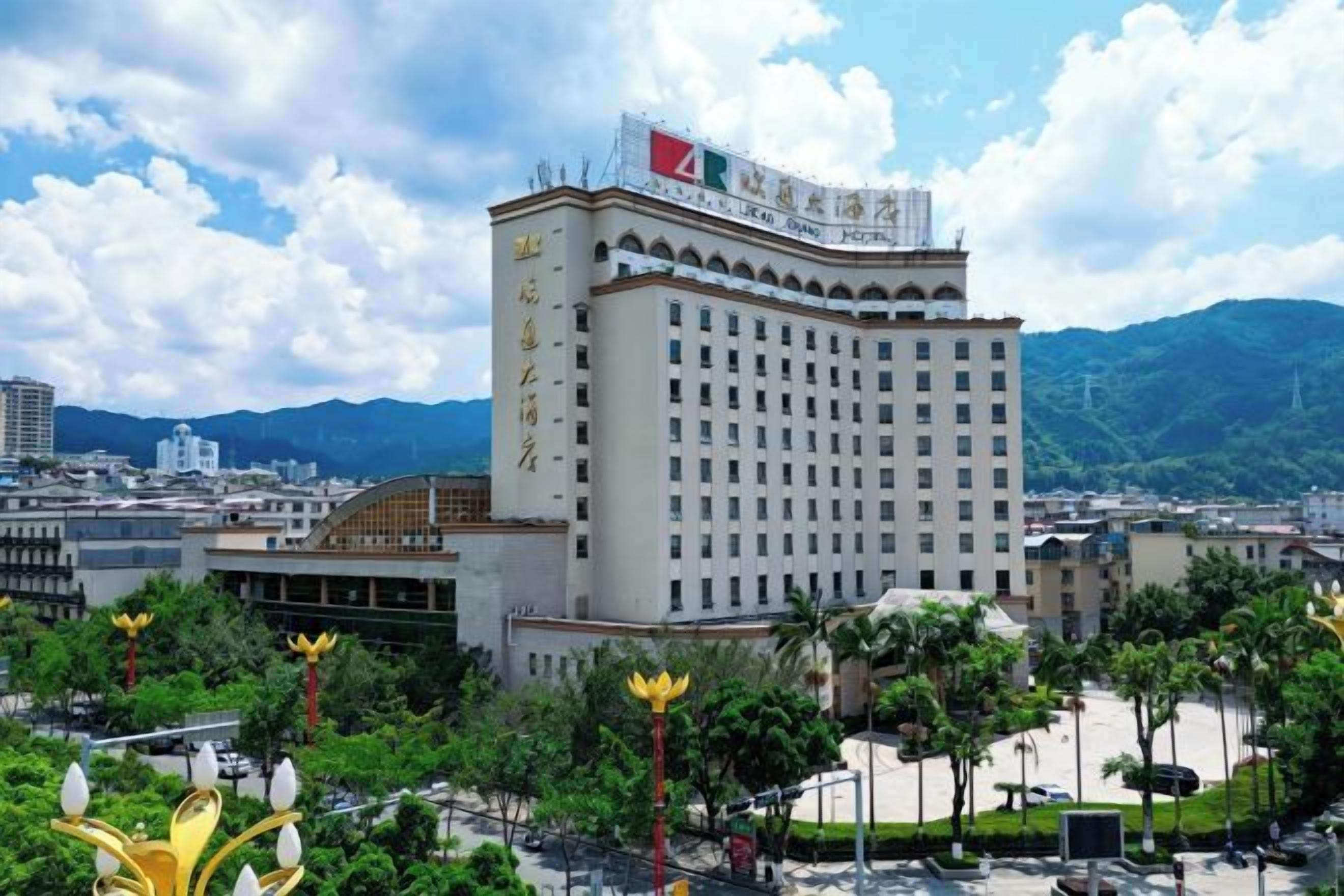 Un hôtel à plusieurs étages, doté d’une enseigne bien visible, entouré de verdure et avec une chaîne de montagnes en arrière-plan.
