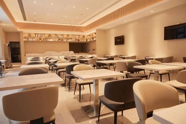 A modern hotel dining area with multiple tables and chairs, a central seating area, and a wall-mounted television.