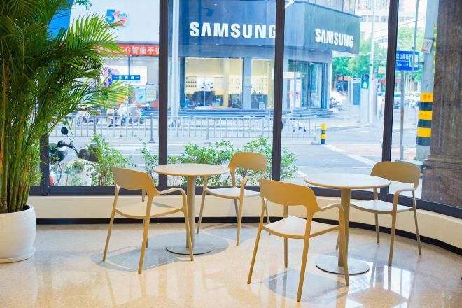 A seating area with tables and chairs, a large window with a view of a street and a Samsung store, and a potted plant.