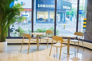 Una zona de estar con mesas y sillas, un ventanal con vista a una calle y una tienda Samsung, además de una planta en maceta.