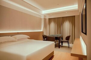 Habitación de hotel con cama, escritorio, dos sillas y un gran ventanal con cortinas.